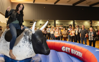 Sarah Toop rides a mechanical bull at the Hometown Hoedown for Hospice while a crowd of smiling guests in western-inspired outfits watches and cheers inside Evergreen Hall.