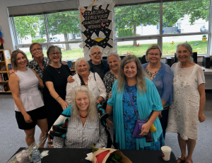 A group of adults encircle Lucy Fraser seated at a table in a school library with large windows behind them.