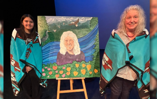 Two people stand on either side of a painted portrait displayed on an easel on a stage with blue lighting.