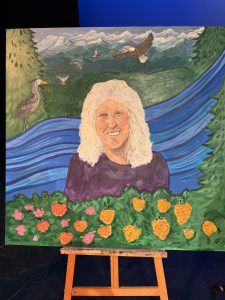 The portrait depicts an older woman against a landscape of mountains, a winding river, birds in flight and colourful flowers, reflecting elements of nature and community.