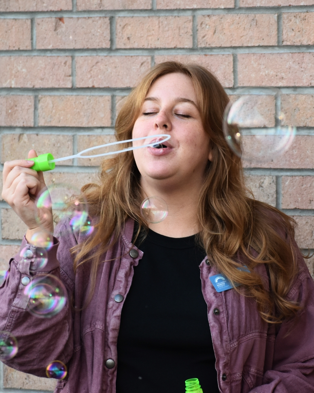 Karlee Gross Chilliwack Hospice Society Child and Youth Program Karlee Gross, Child and Youth Program Manager at Chilliwack Hospice Society, blows bubbles outside in front of a brick wall.